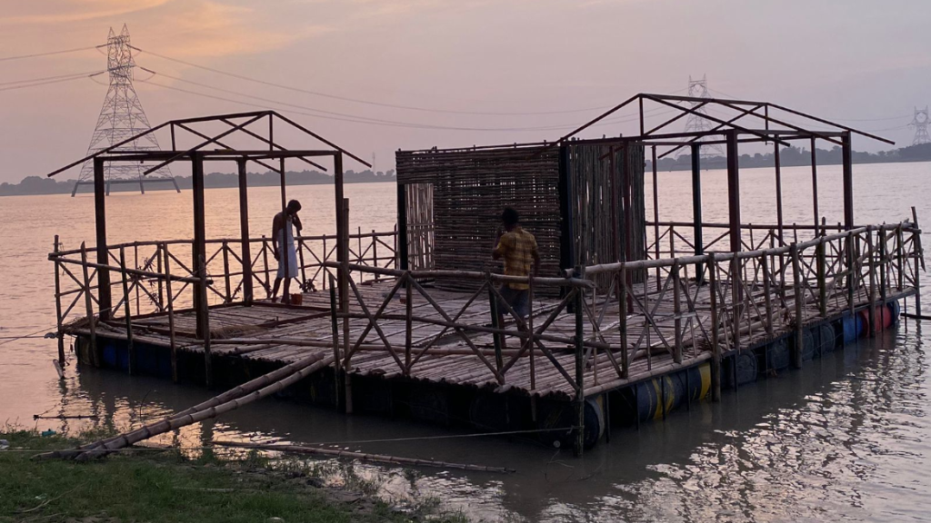 Two individuals are diligently working on the City App prototype, a floating structure under construction on the Ganges river in Bihar. The structure, built upon barrels for buoyancy, is divided into three sections. One section is already covered with wooden planks, the two other sections have incomplete wooden frames, indicating ongoing construction. The soft lighting of dusk bathes the scene, highlighting the intricate details of the structure and the calm waters around it. In the distance, an electricity pylon and transmission lines stand against the evening sky, adding a touch of modernity to this serene riverscape