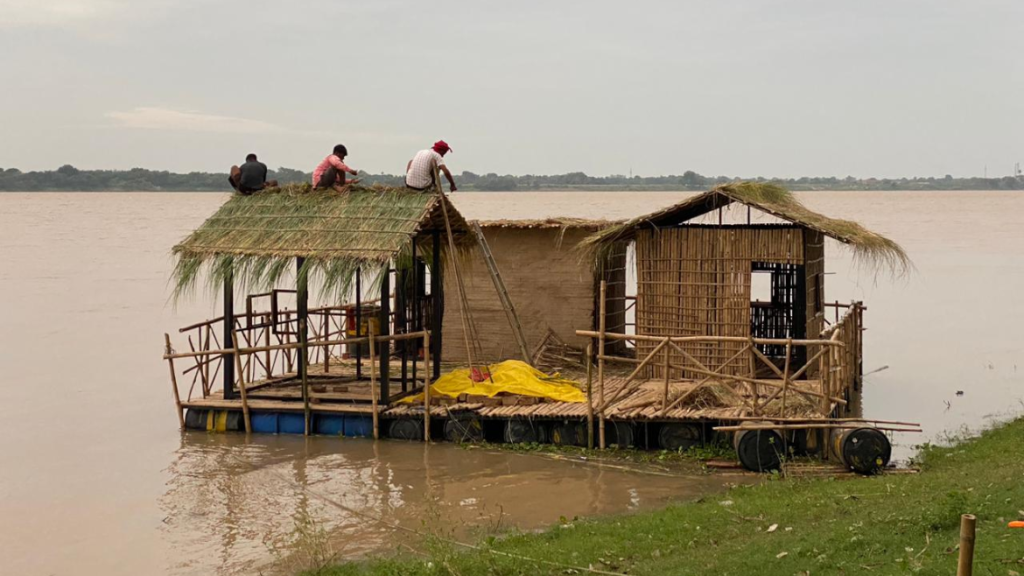 A group of three people is working on the thatched roof of the City App prototype, in Bihar. This floating structure is moored to the grassy shores of the Gange, under an overcast sky.