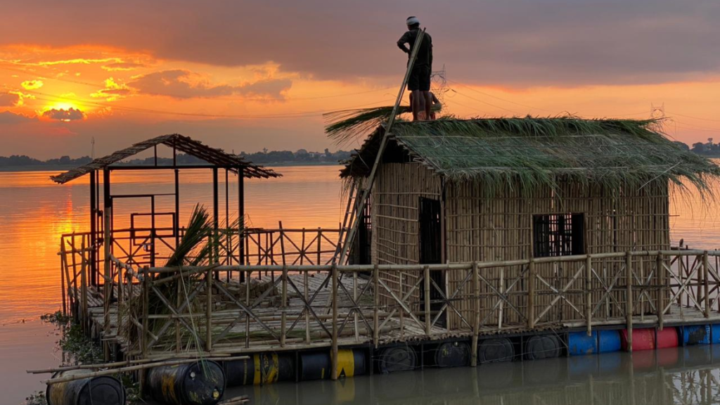 Under a sky awash with the sun’s golden and crimson hues, a solitary figure is perched on a floating bamboo structure, the City App prototype under construction. Bathed in the sunset’s glow, the structure stands out against the backdrop of dancing tree silhouettes and other elements of nature.